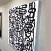 Robert R – Black and White Modern Abstract Painting, Angled Detail Close-up angled view of a black-and-white modern abstract painting with bold graffiti-inspired lines and a visible white canvas edge against a gallery wall.